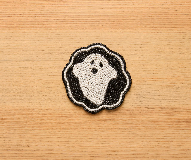 Handmade round beaded coaster with a black background and a white smiling ghost design, crafted from glass beads and finished with a scalloped edge.