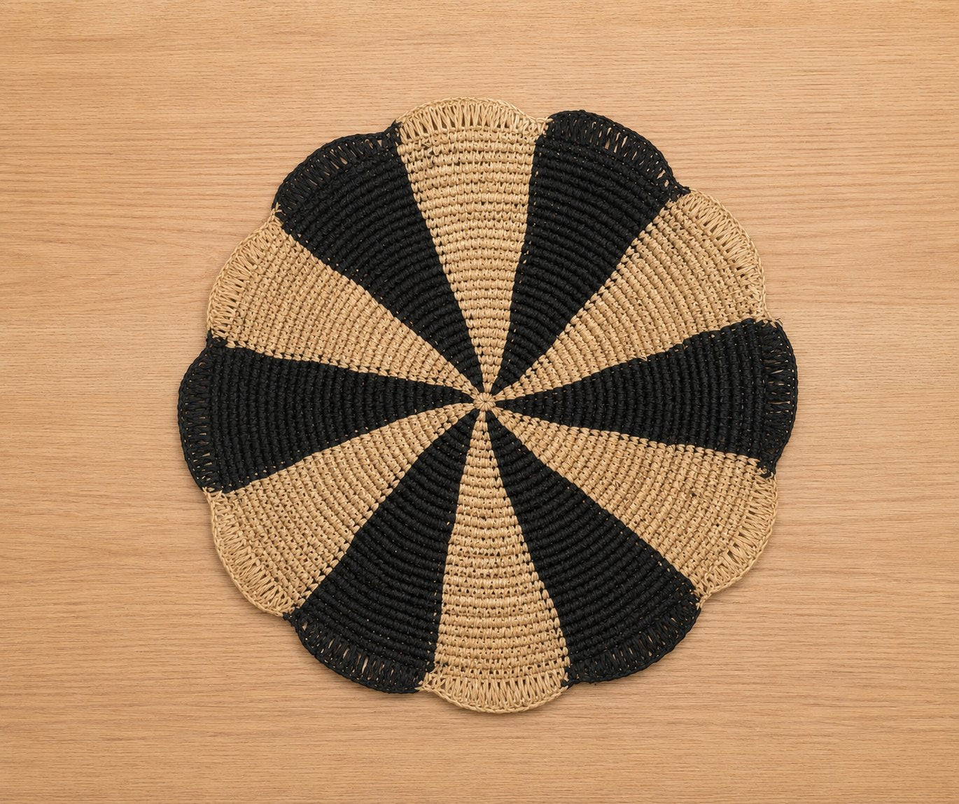 Round scalloped raffia placemat with black and natural pinwheel pattern on a light wood table