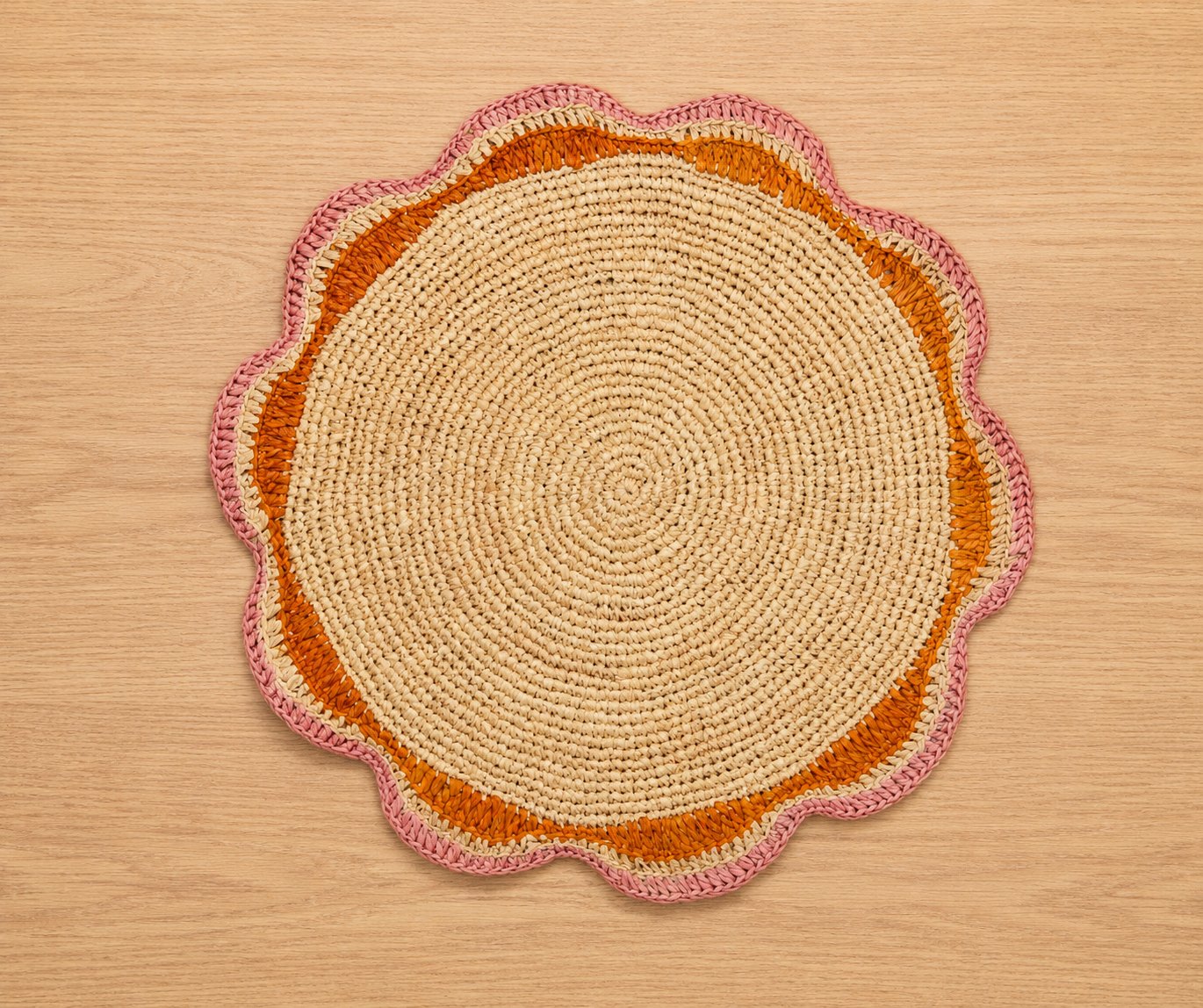 Round scalloped raffia placemat with pink and orange border on a light wood table
